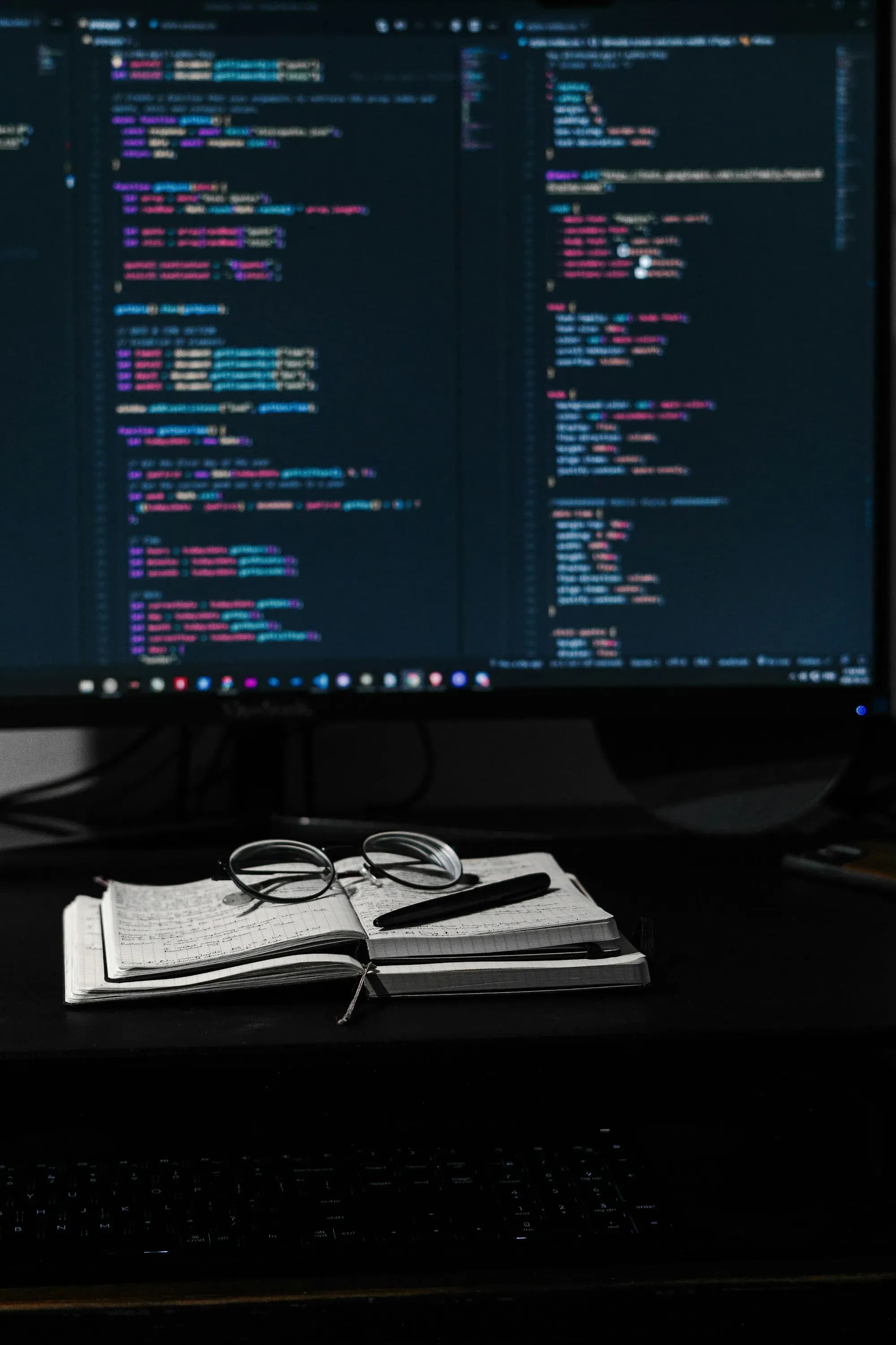 Zwei Monitore mit Quellcode, davor ein Notizbuch mit Brille